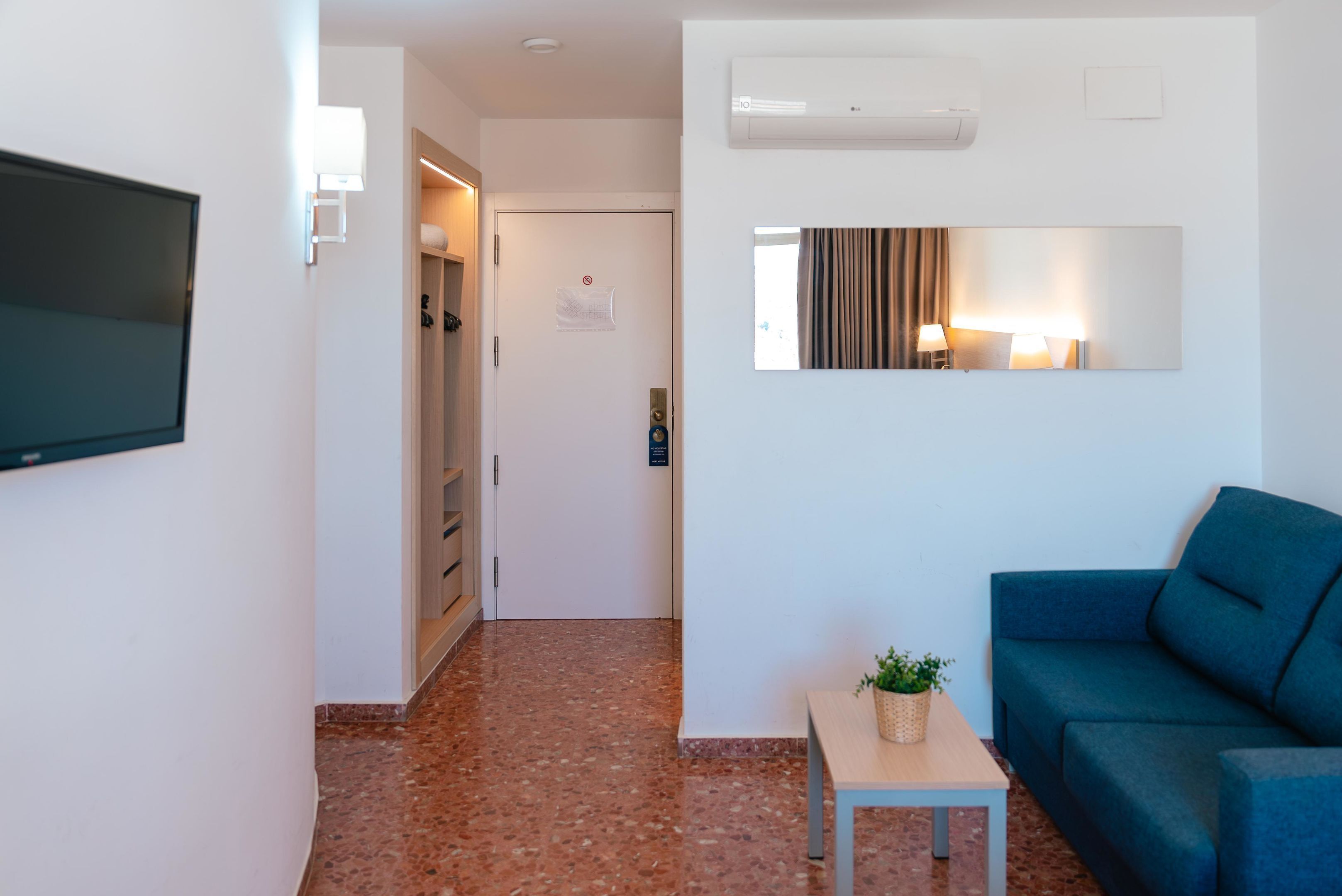 Habitación de hotel moderna con televisor de pantalla plana, una pequeña mesa con una planta, sofá azul, espejo y puerta que da a un pasillo.