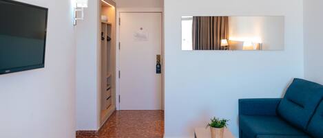 A modern hotel room with a flat-screen TV, a small table with a plant, a blue sofa, a mirror, and a door leading to a hallway.