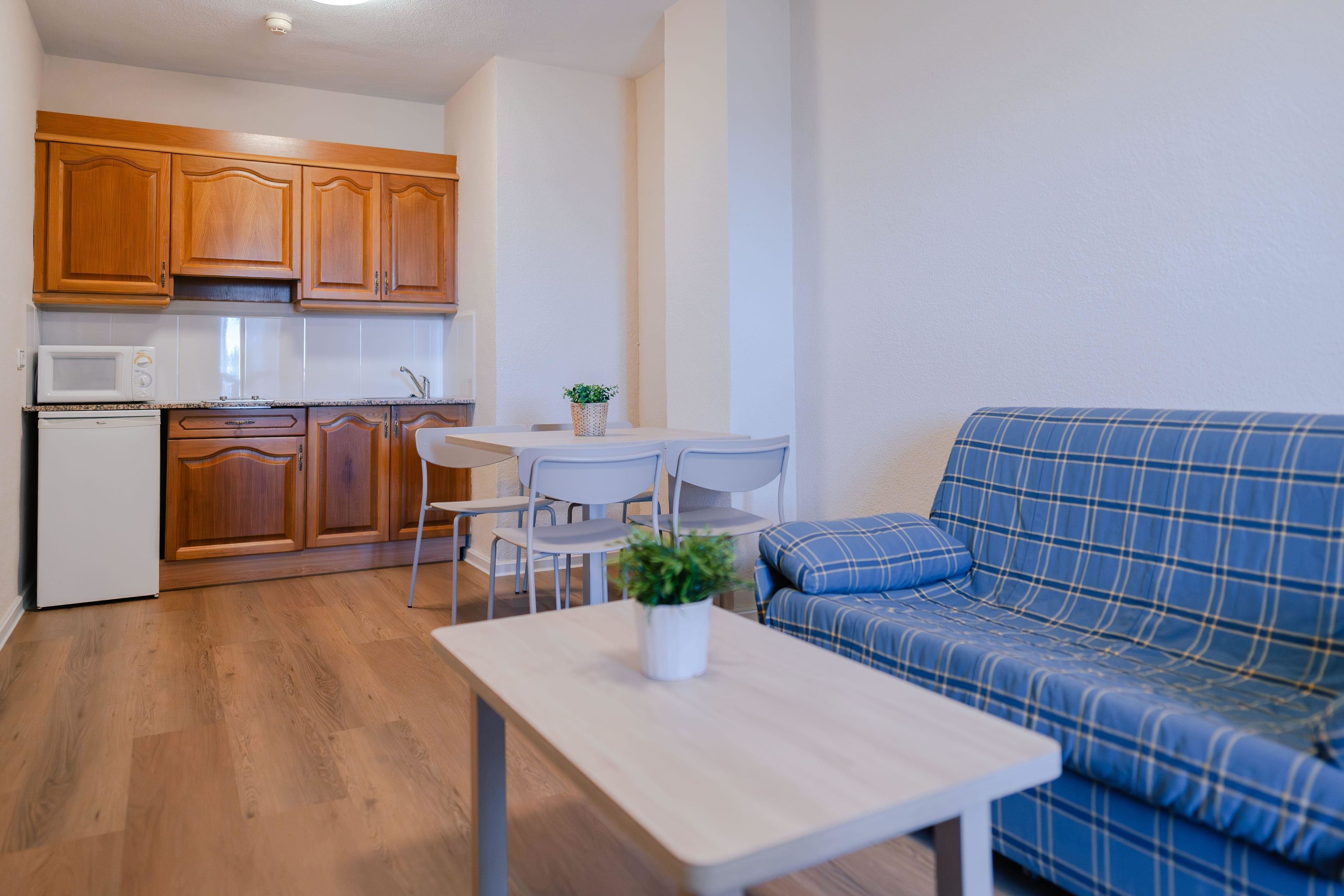 A compact living space with wooden cabinets, a microwave, a dining table with chairs, and a blue plaid sofa.