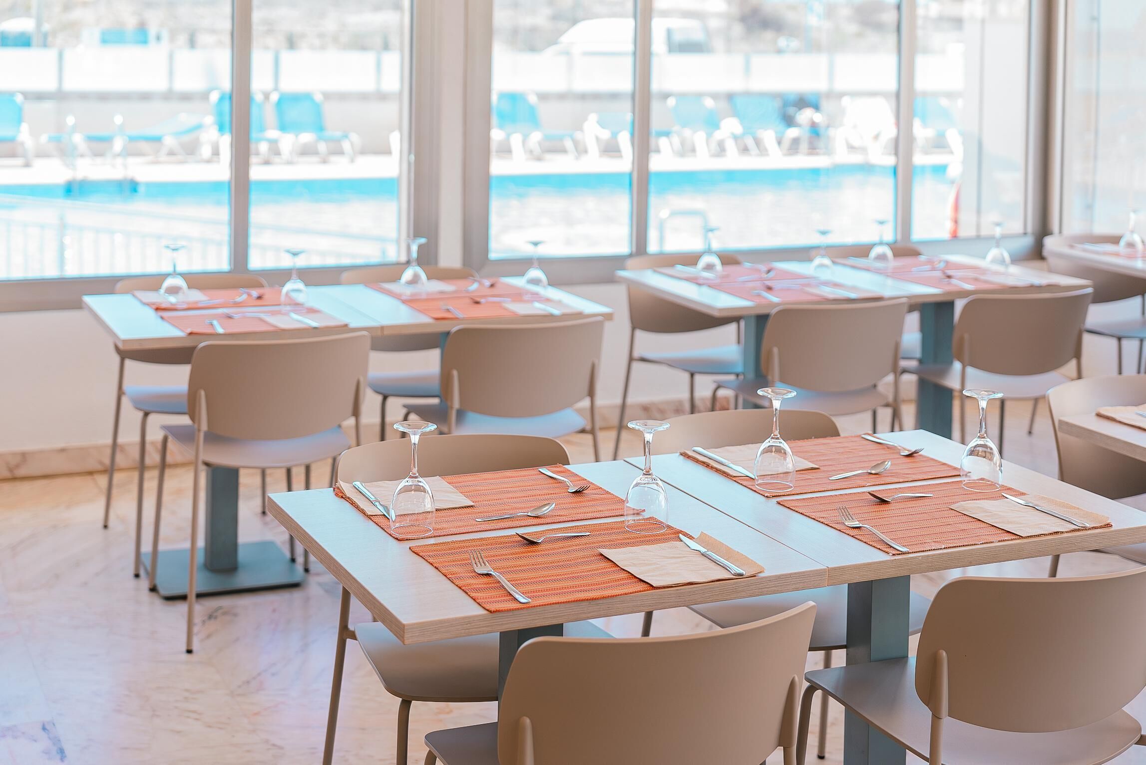 A restaurant with tables set for dining, chairs, and a view of a pool through large windows.