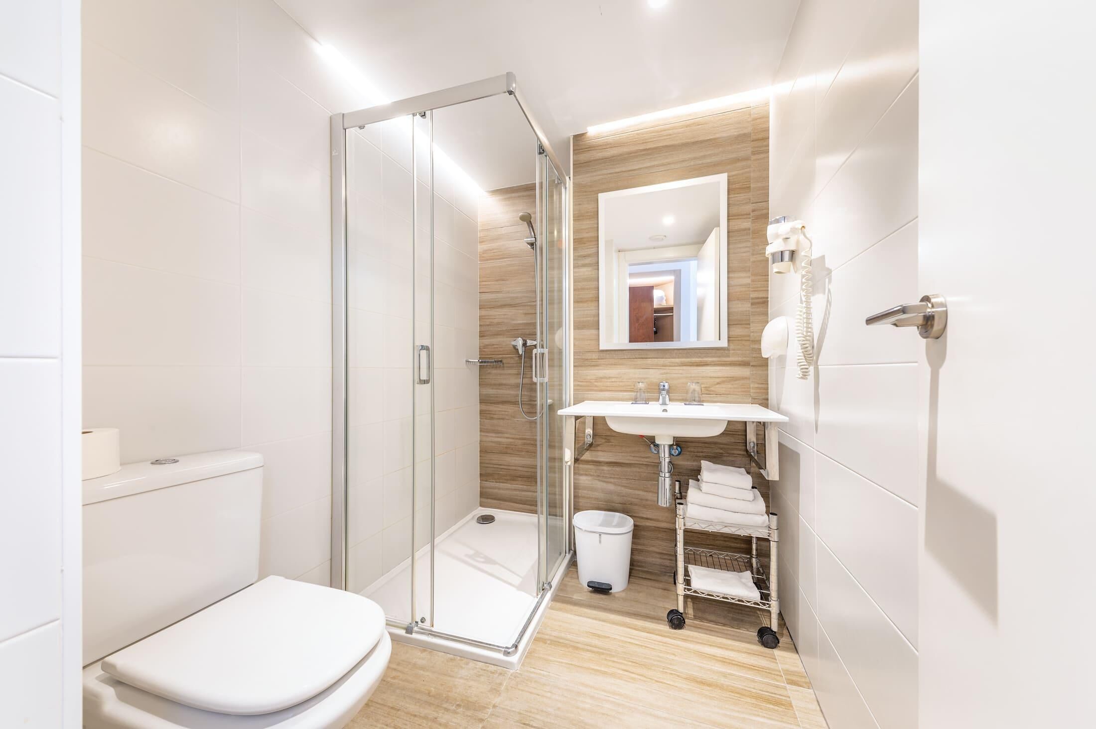 Un baño moderno con ducha de vidrio, inodoro blanco, lavamanos con espejo y toallero.