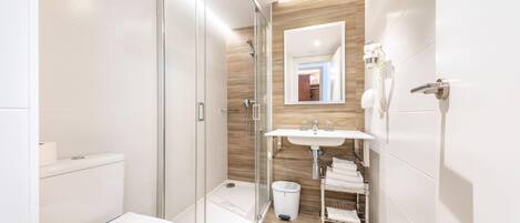 Un baño moderno con ducha de vidrio, inodoro blanco, lavamanos con espejo y toallero.