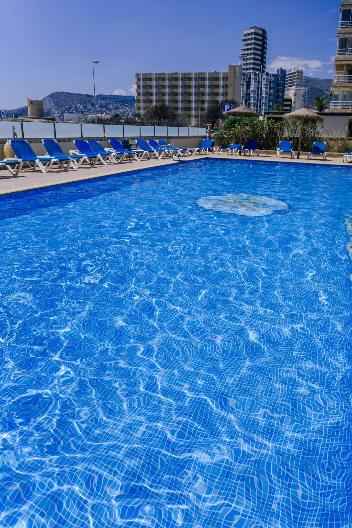 Una piscina con agua azul, rodeada de sillas de descanso y sombrillas azules, con edificios al fondo.