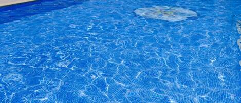 Una piscina con agua azul, rodeada de sillas de descanso y sombrillas azules, con edificios al fondo.