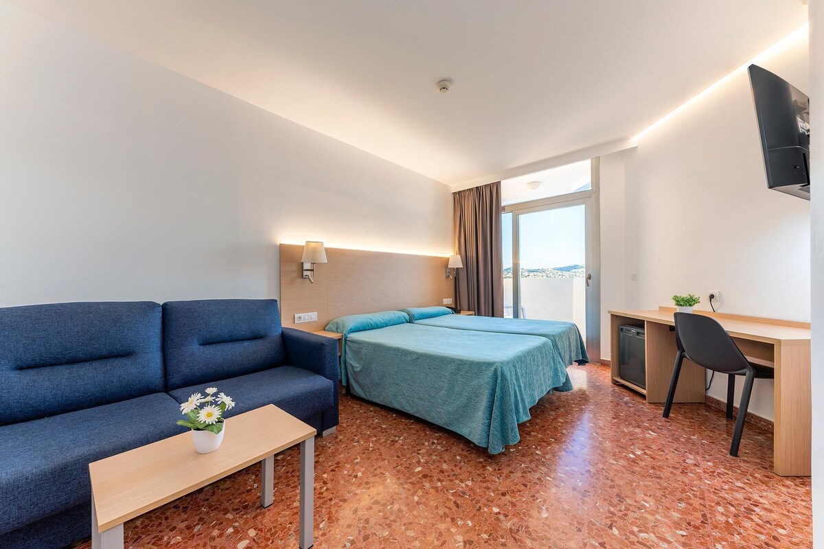 Habitación de hotel con sofá, dos camas, escritorio, una silla y una mesita con un florero.