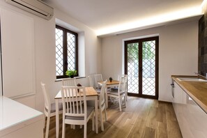 A modern kitchen with a dining area, featuring a wooden table and white chairs, large windows with metal grilles, and a view of greenery outside. - Maria Rosaria Bed & Breakfast (Ladispoli)