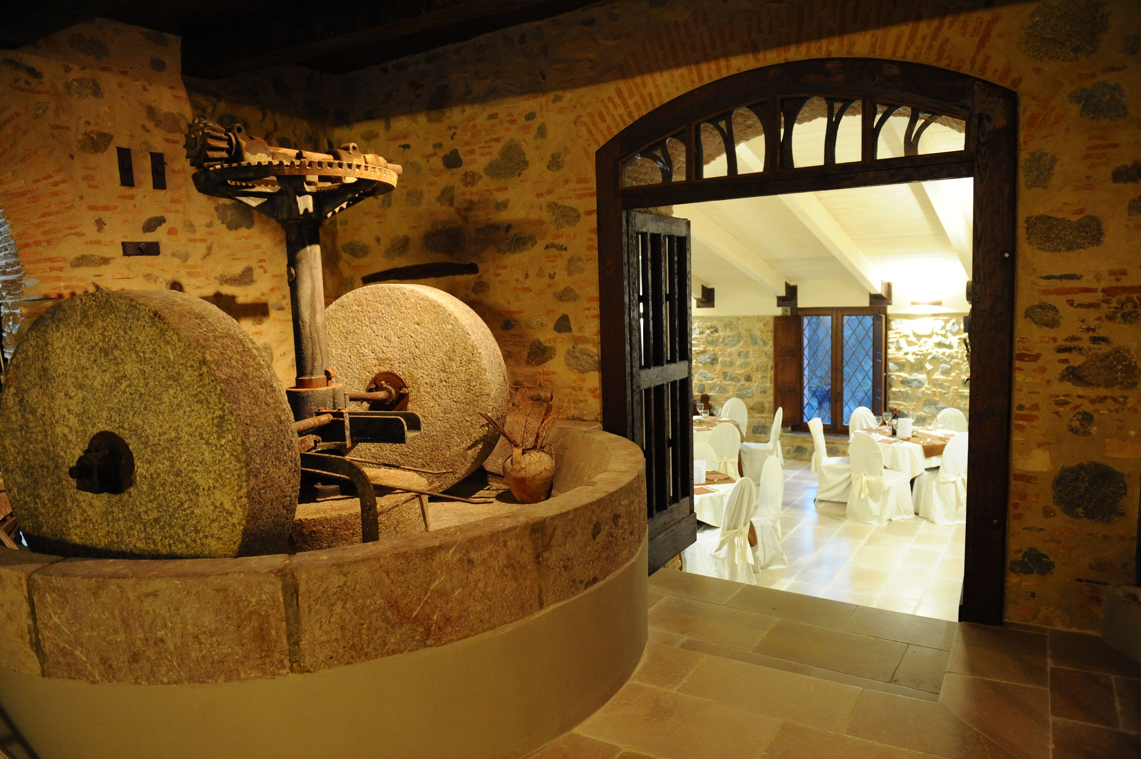 Un espacio con suelo de piedra, una gran pila de piedra, una estructura de madera y un arco que da a un comedor con manteles blancos y sillas.