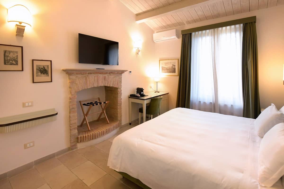 A hotel room with a bed, a desk with a chair, a fireplace, a television mounted on the wall, and a window with curtains.