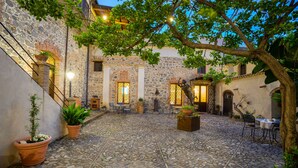 A courtyard with stone flooring, potted plants, outdoor seating, and a stone building with arched windows.