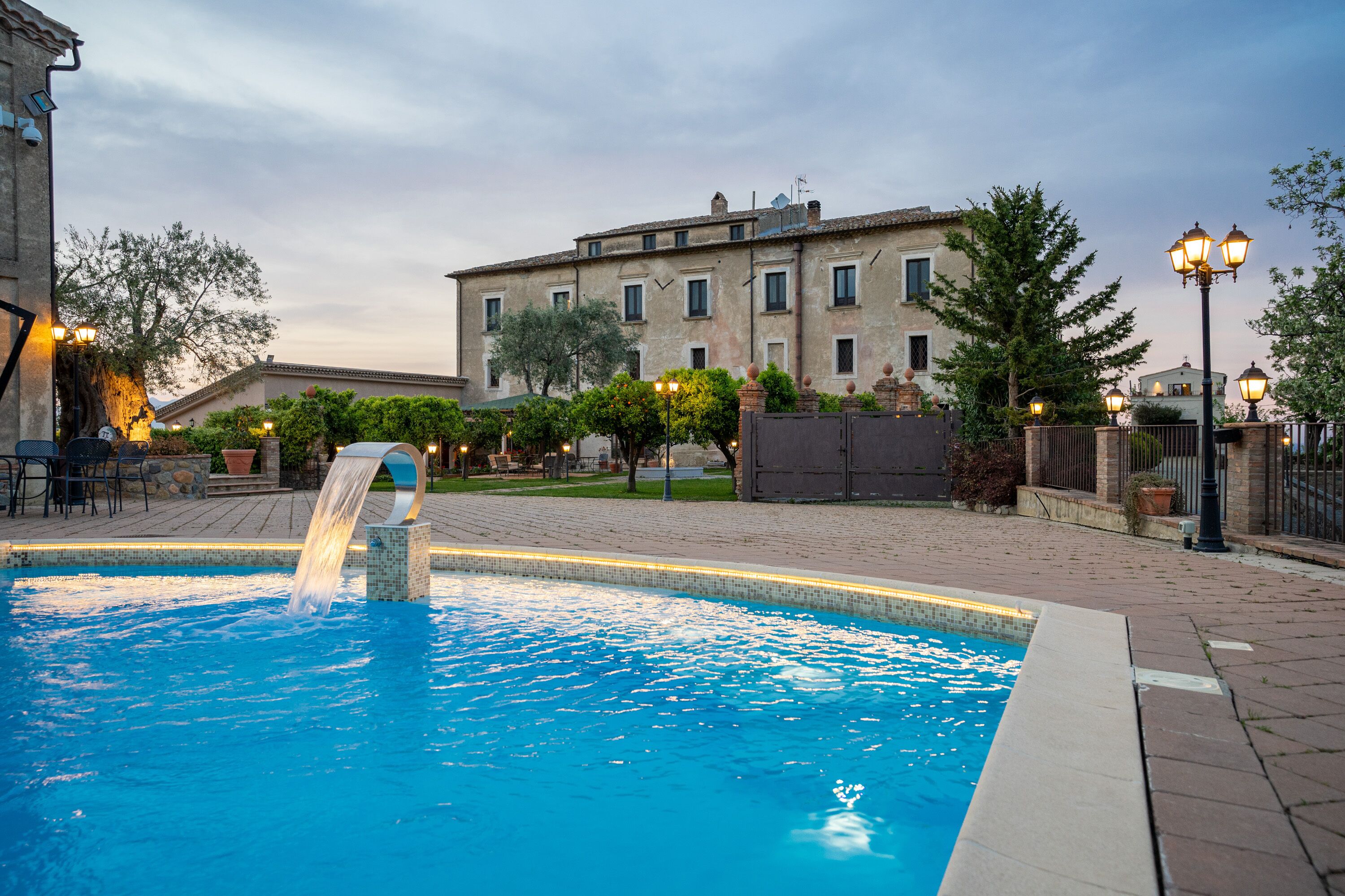 Una piscina con cascada, rodeada por un patio de ladrillo y un edificio con varias ventanas.