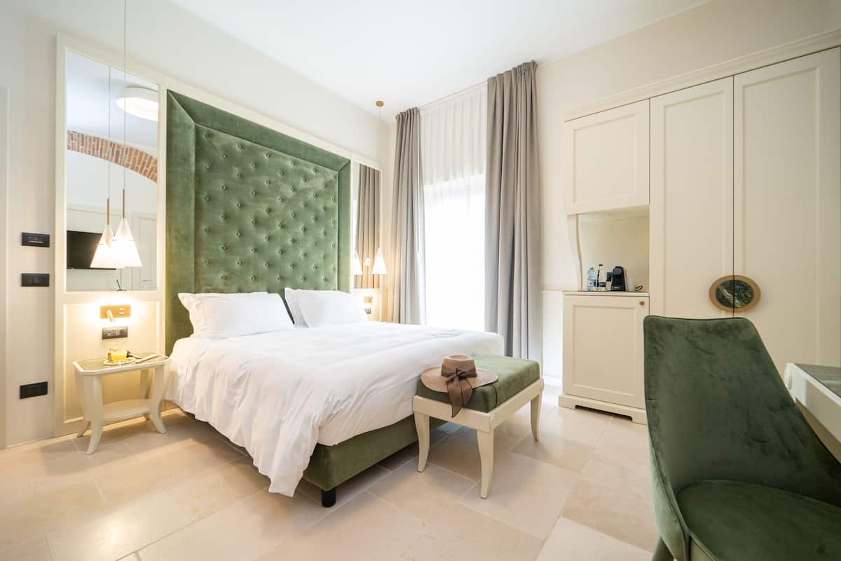 A hotel room with a large bed, a green upholstered headboard, a bedside table with a lamp, a small stool with a hat, and a green armchair.
