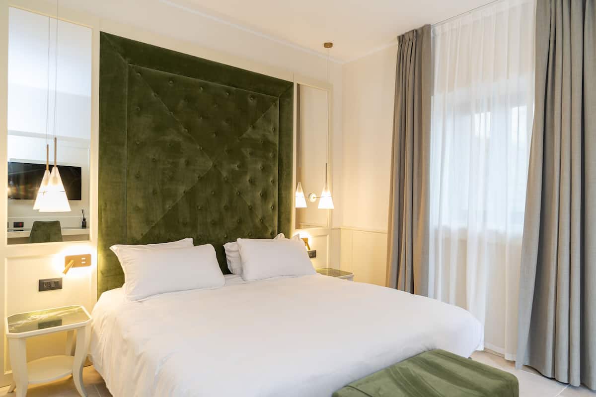 A hotel room with a large bed, a green upholstered headboard, bedside lamps, a small table, and a view of a window with curtains.