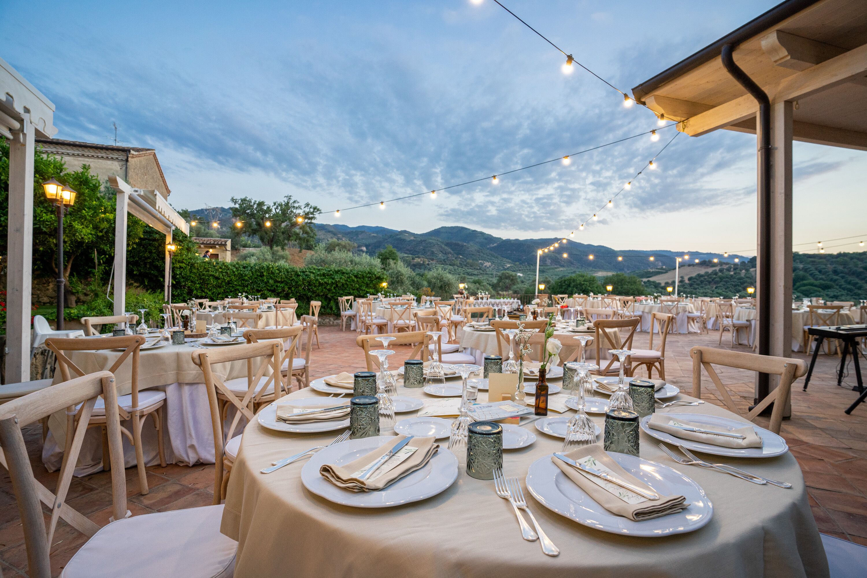 An outdoor dining area with round tables set for a meal, surrounded by chairs, and string lights overhead.