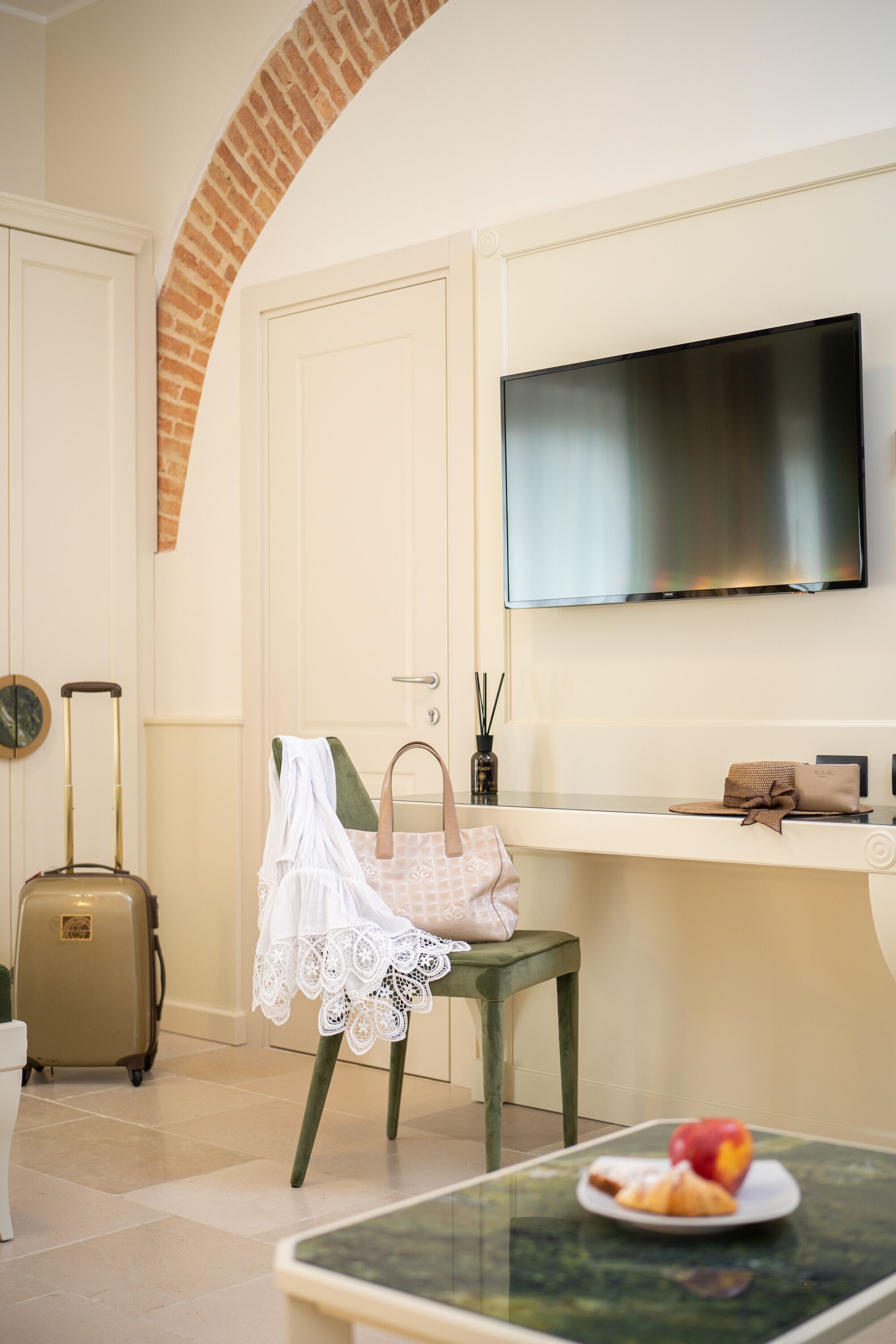 Habitación de hotel con un televisor montado en la pared, una maleta, una silla con cubierta de encaje blanco y una mesa con un plato de comida.