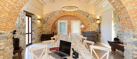 Una larga mesa con una computadora portátil, un cuaderno y un portalápices en una habitación con paredes de piedra y vigas de madera.