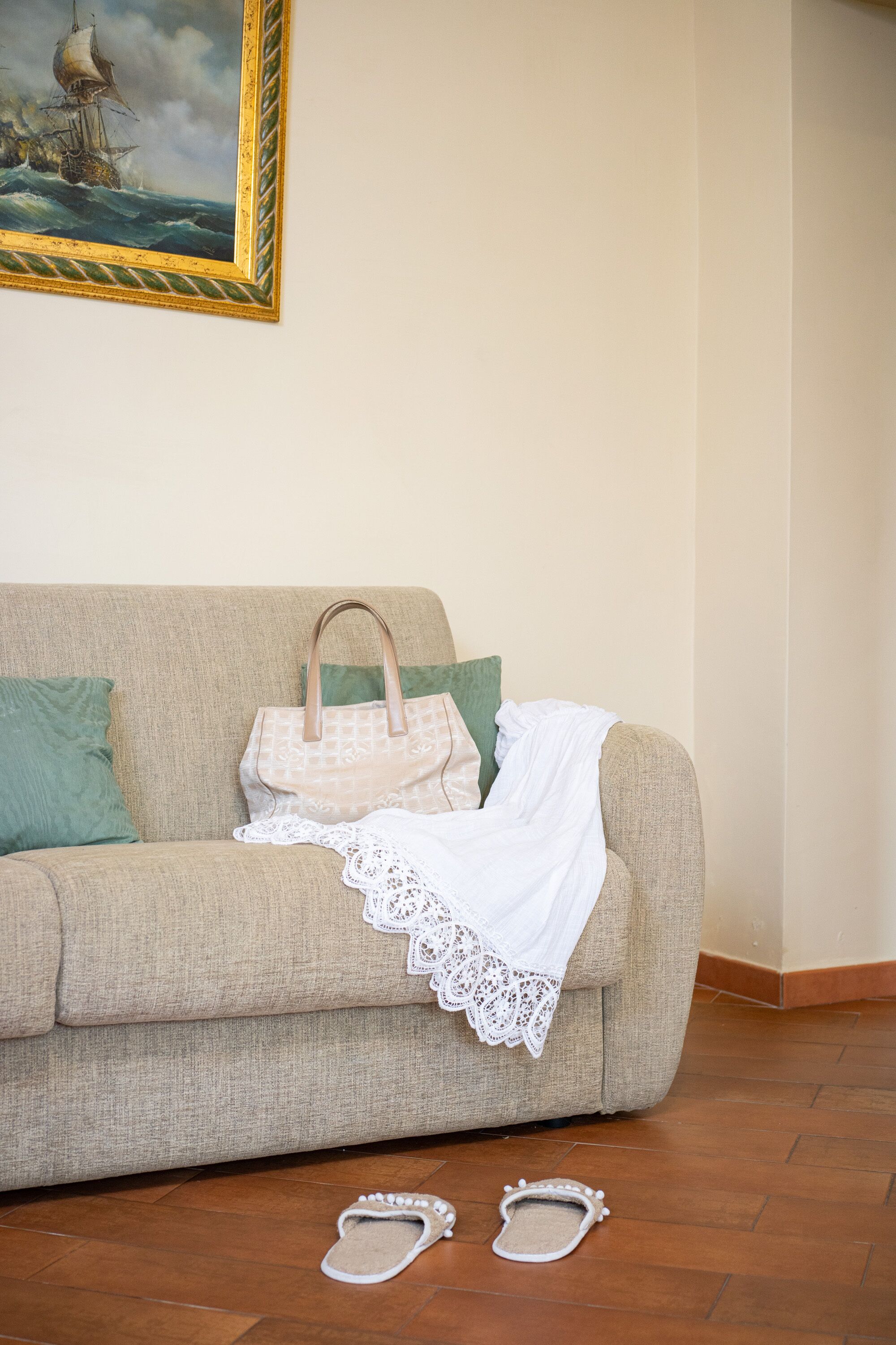 Un canapé beige surmonté d’un jeté de dentelle blanche, un sac à main beige et une paire de chaussons sur un plancher en bois.
