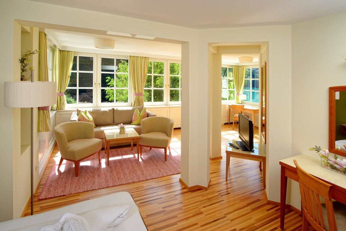 Habitación luminosa con sofá, dos sillones, una mesita, televisor y escritorio con espejo. Amplias ventanas con cortinas dejan entrar luz natural.