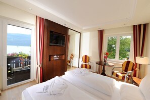 Habitación de hotel con cama, televisor, escritorio con silla, ventana con cortinas y balcón con flores y vista al mar.