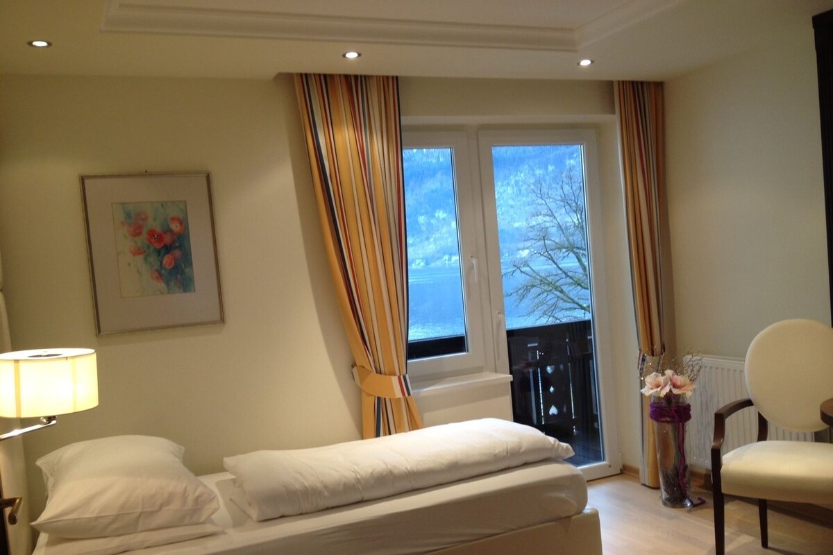 Habitación de hotel con cama, una silla, una lámpara, un cuadro y una ventana con cortinas.