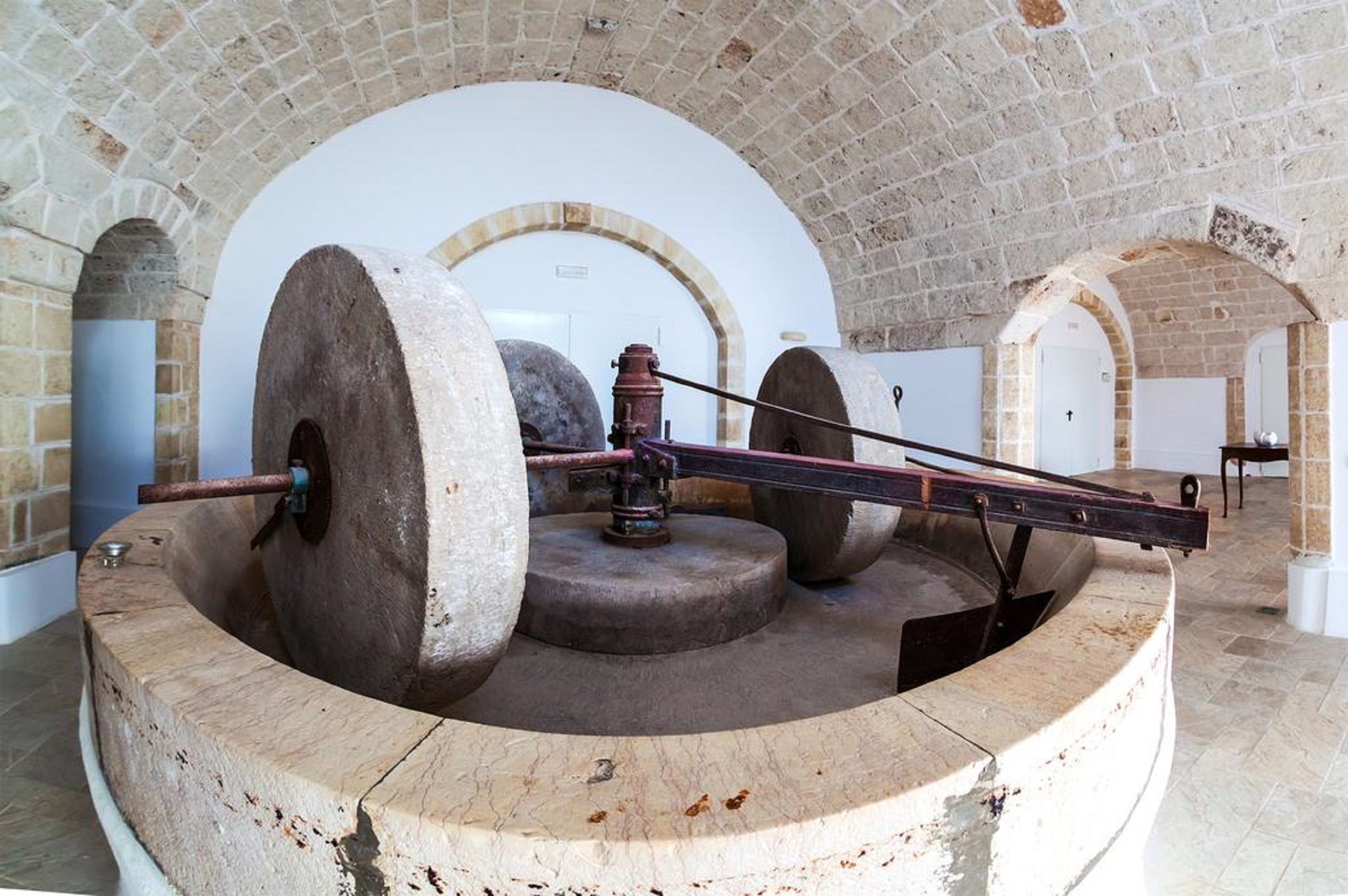 Un mécanisme historique de roue à eau, doté de grandes roues en pierre et d’une poutre métallique, installé dans une pièce aux murs de pierre et aux plafonds en arc de cercle.