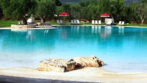 Ein Swimmingpool mit Liegestühlen und Sonnenschirmen, umgeben von Bäumen und einer Steinstruktur.