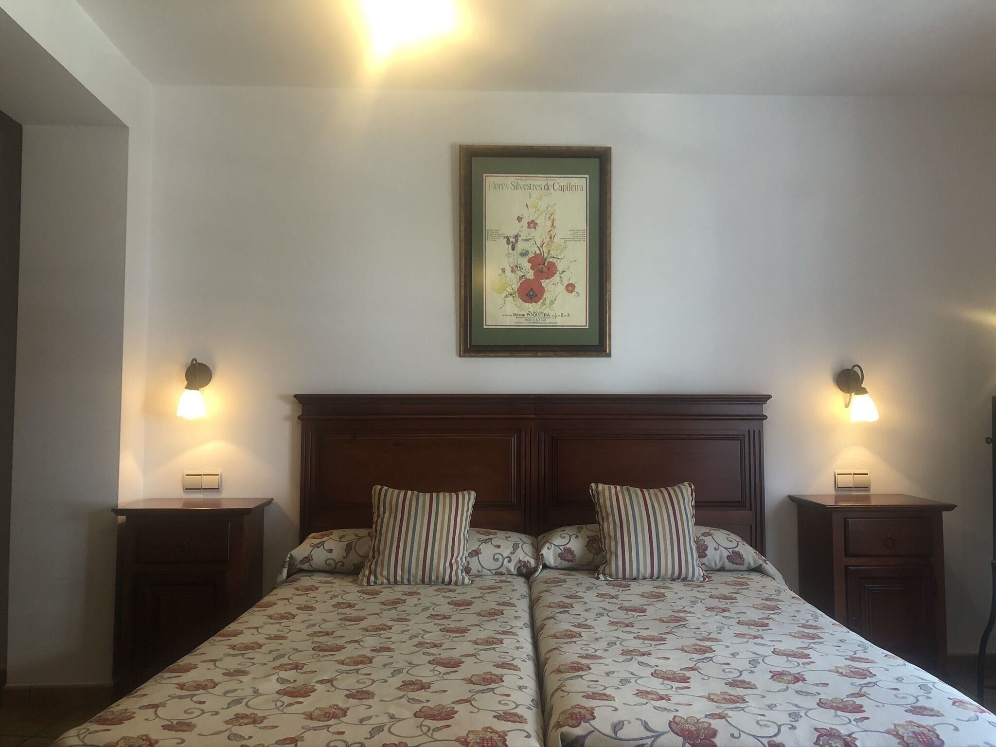 A double bed with floral bedding, two bedside tables, a framed botanical illustration, and wall-mounted lamps.