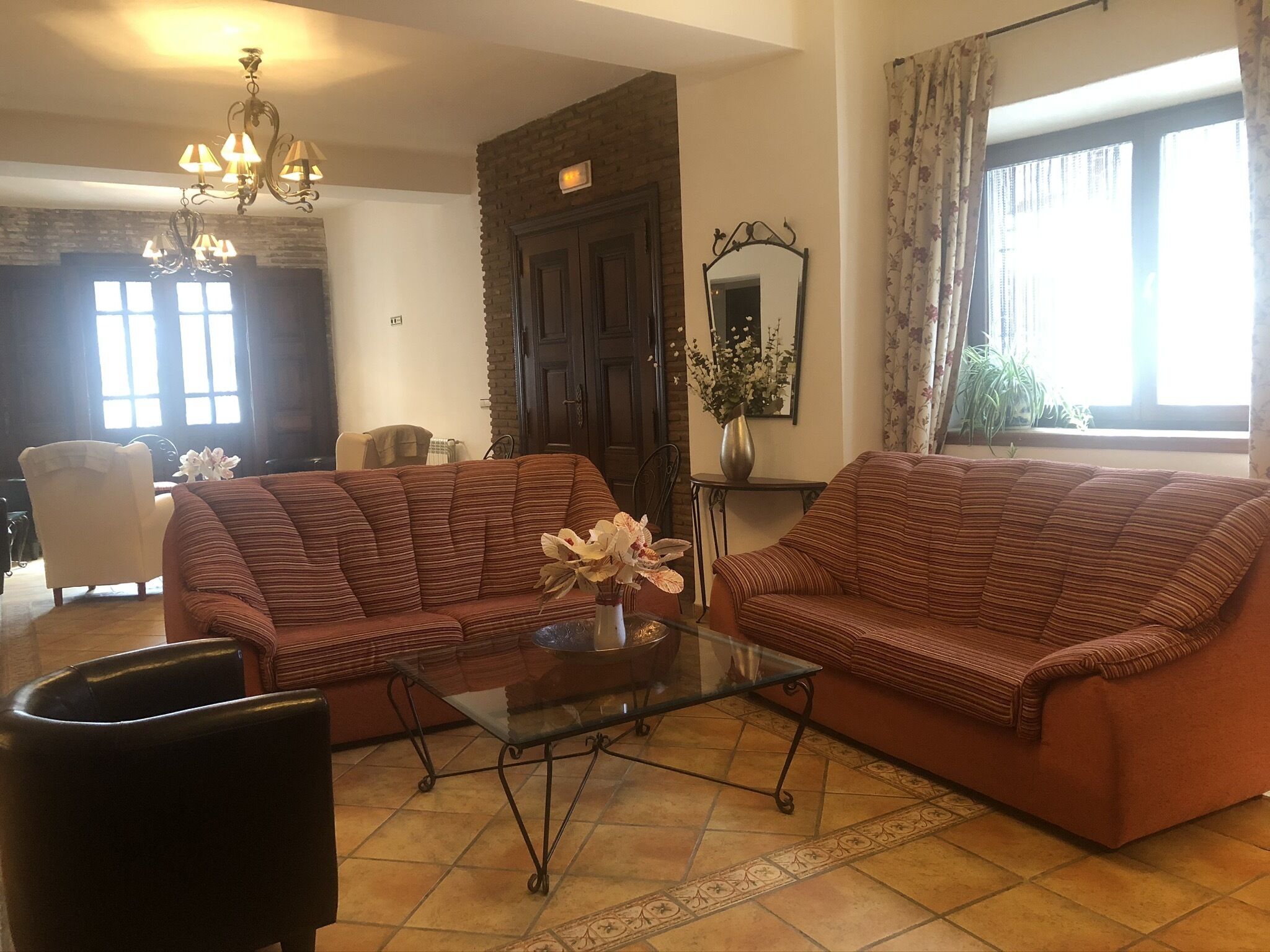 A living room with two striped sofas, a glass coffee table, a white armchair, a chandelier, and a large window with curtains.