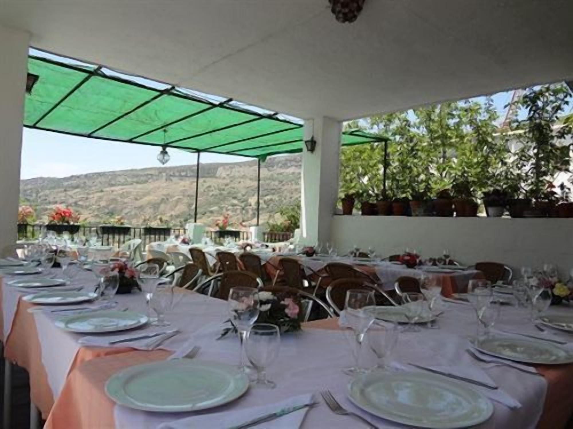 A covered outdoor dining area with tables set for a meal, potted plants, and a view of the hills.