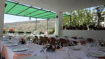 A covered outdoor dining area with tables set for a meal, potted plants, and a view of the hills.