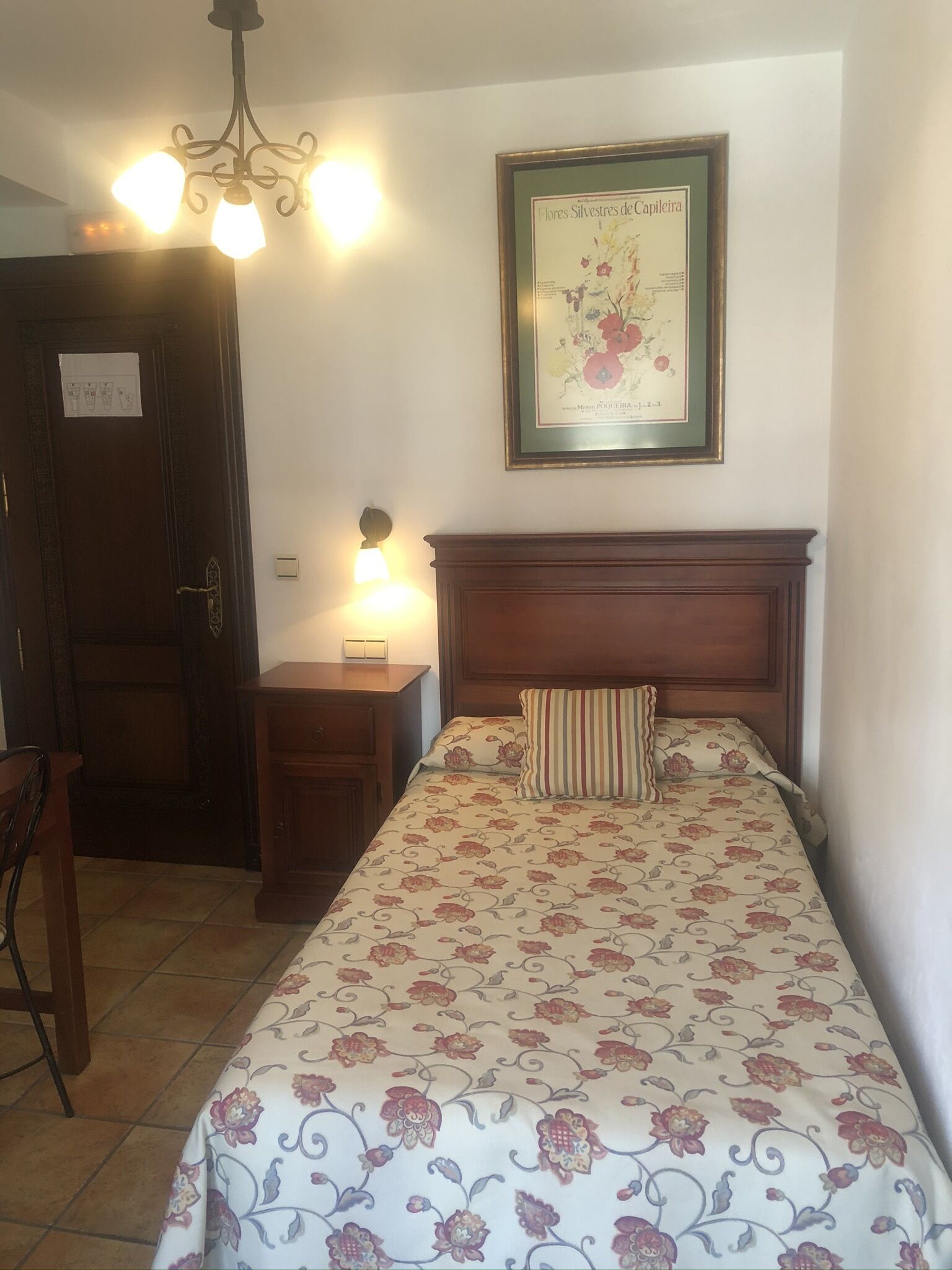 Un dormitorio con una cama, una mesita de noche, una silla, una puerta de madera y una lámpara fijada a la pared.