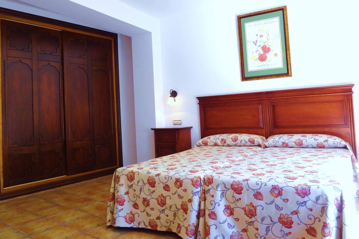 Un dormitorio con cama de madera, colcha floral, una mesita de noche, una lámpara de pared y un cuadro enmarcado.