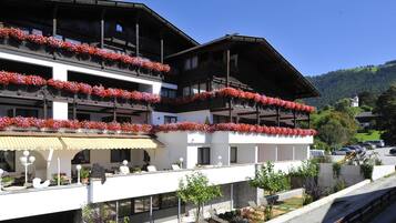 Un hotel con varios balcones decorados con flores rojas, mobiliario exterior y vistas a una ladera verde.