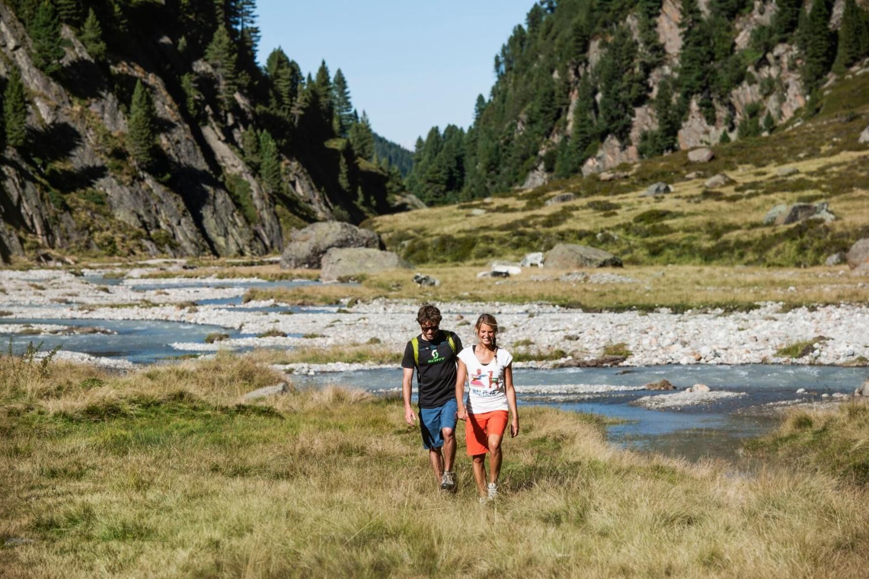 A couple walking along a riverbank with a rocky landscape and trees in the background.