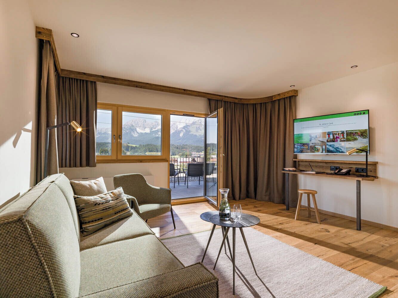 Una sala de estar moderna con un sofá, una mesa de centro, un televisor y vistas a montañas a través de amplios ventanales.
