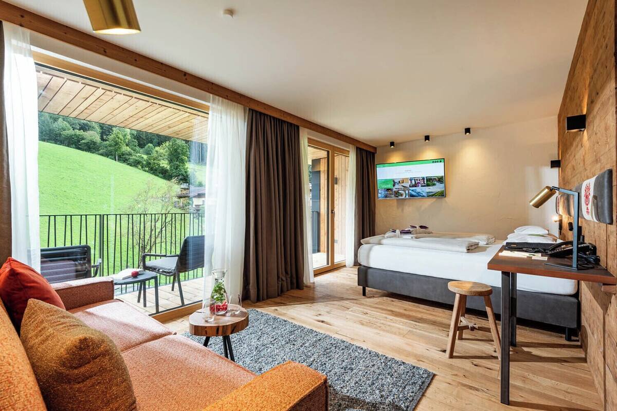 A modern hotel room with a bed, a desk, a sofa, and a balcony with a view of a green landscape.