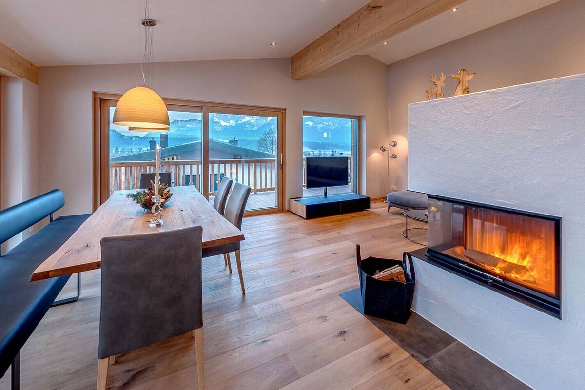 A modern living room with a wooden dining table, chairs, a sofa, a fireplace, and large windows offering a mountain view.