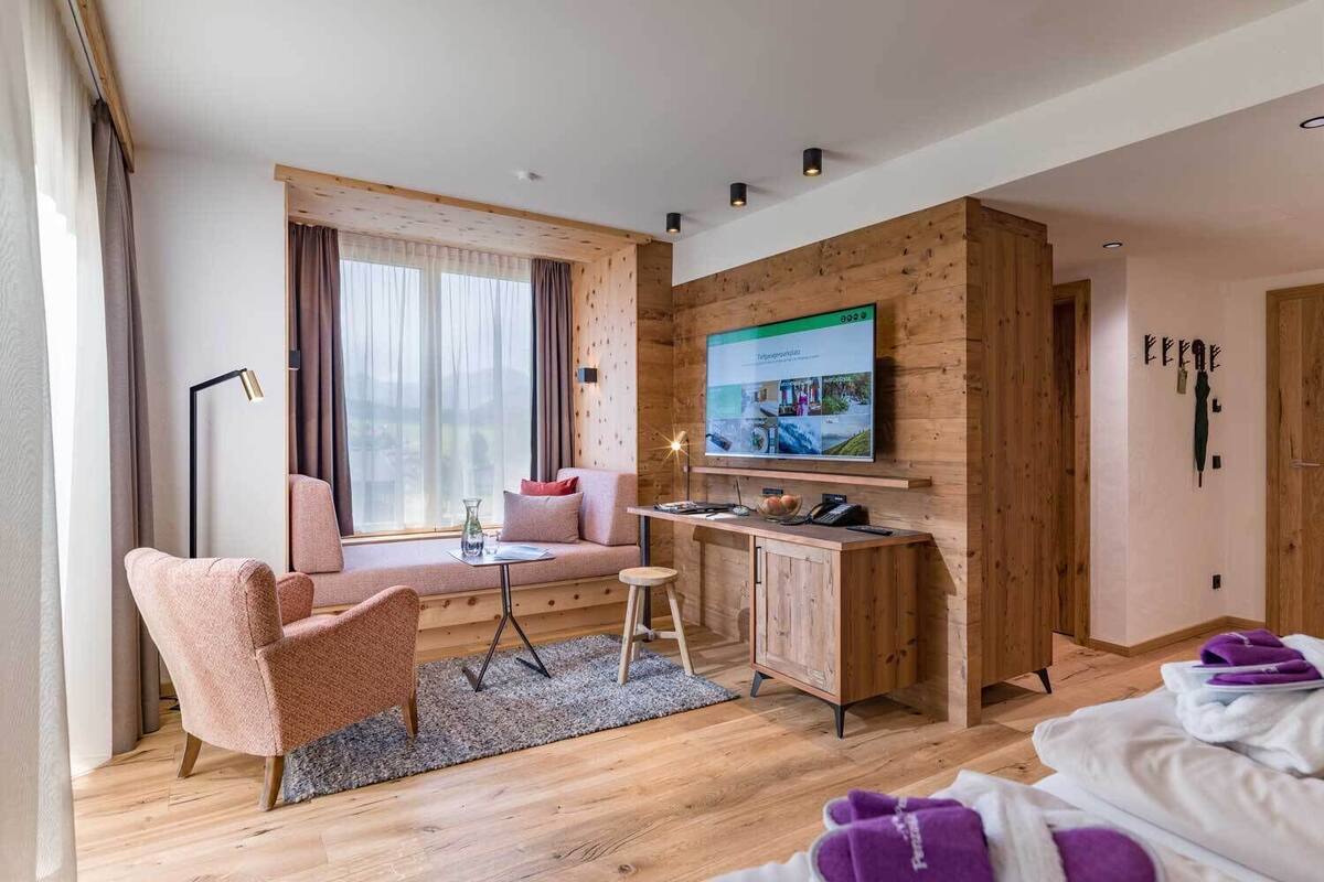 A modern hotel room with a wooden accent wall, a large flat-screen TV, a sofa, a desk with a chair, and a bed with a purple blanket.