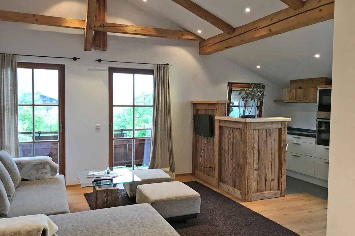 A spacious living area with a sofa, ottoman, and a wooden entertainment unit with a TV. There are large windows and a view of greenery outside.