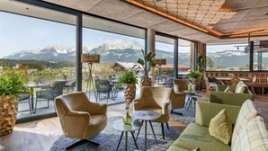 Un interior moderno con un ventanal grande que ofrece vistas a la montaña, sillones de cuero, un sofá verde y una pequeña mesa redonda con un florero.