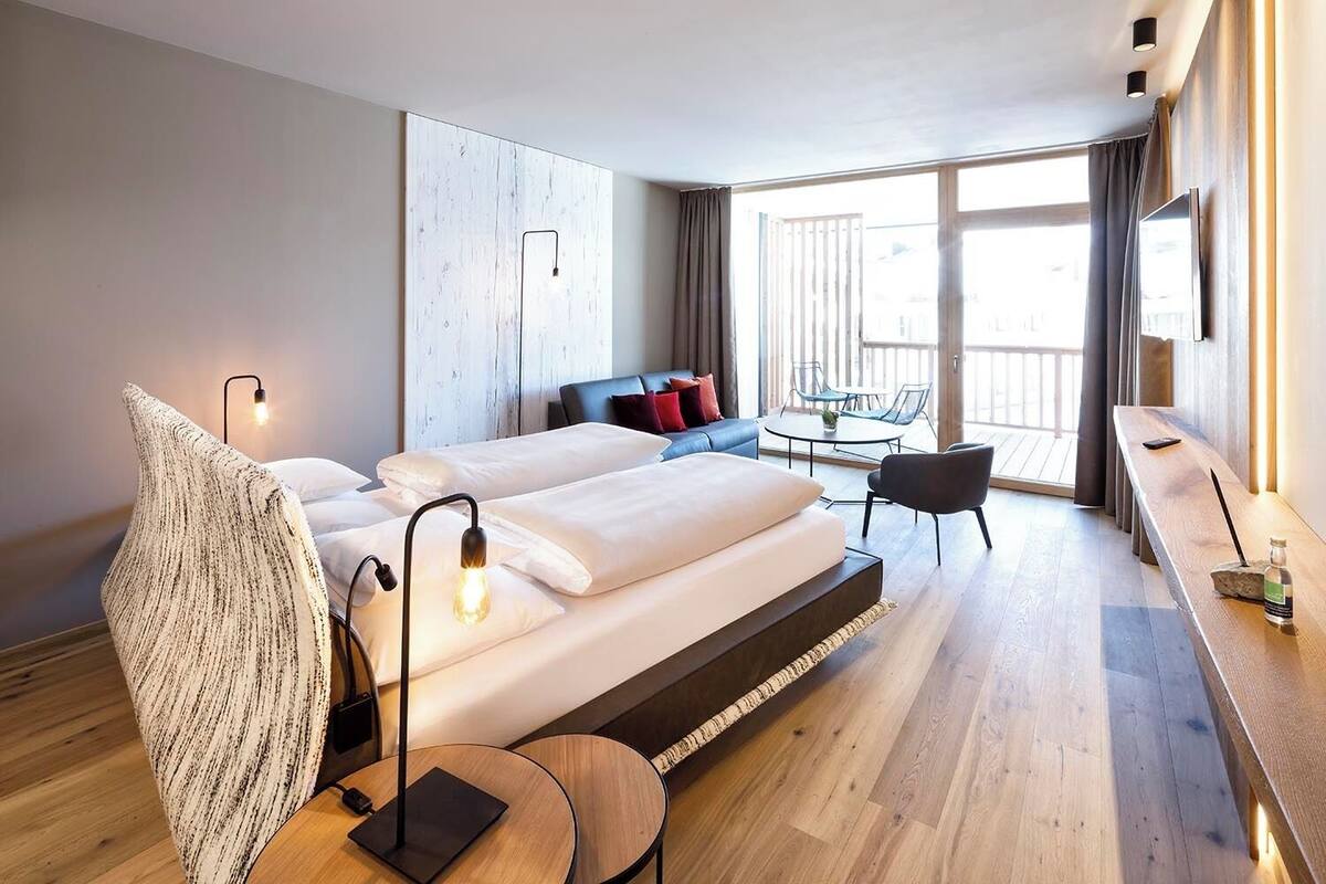 A modern hotel room with a large bed, a desk, a chair, and a balcony with a table and chairs.