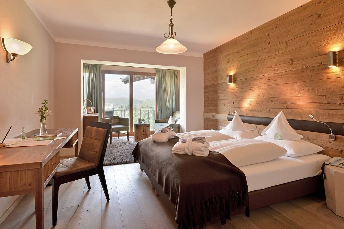 A hotel room with a large bed, a desk with a chair, a balcony with a view, and wooden paneling on the walls.