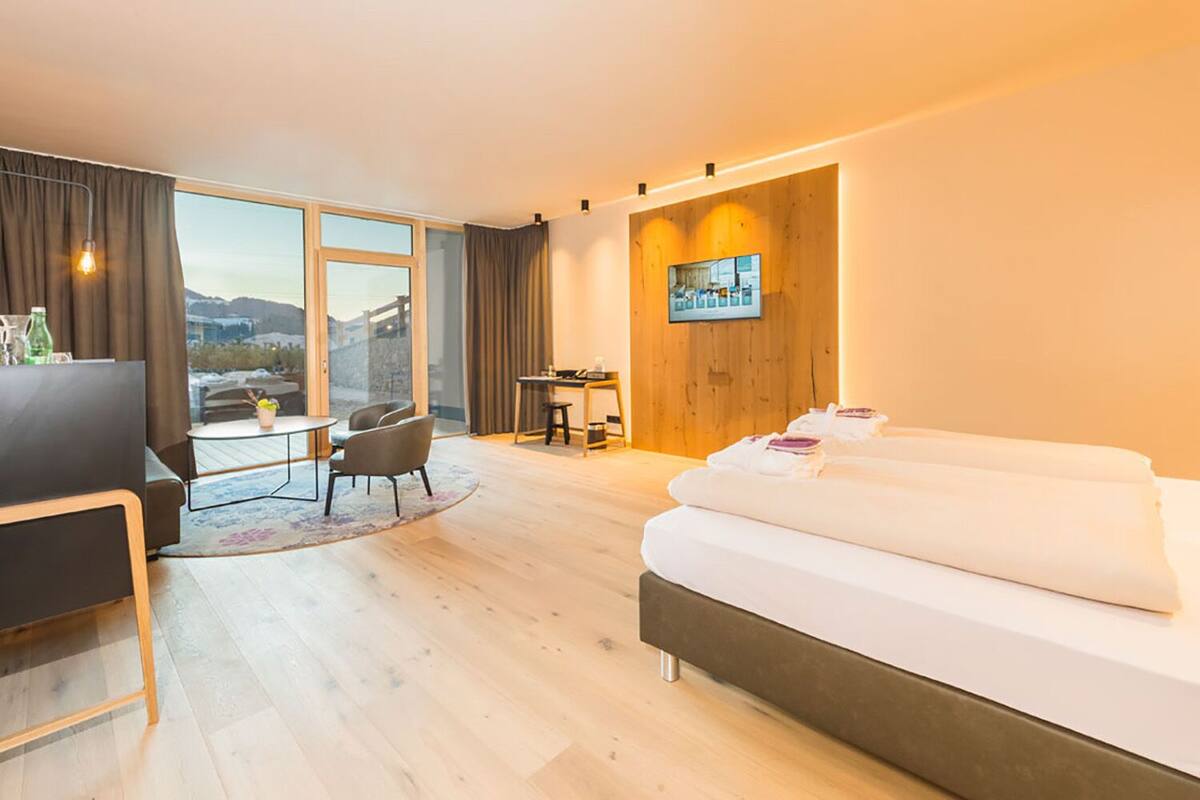 A modern hotel room with a large bed, a desk, a chair, and a view of the mountains through the window.