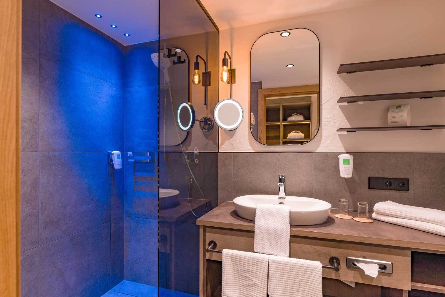 A modern bathroom with a glass shower enclosure, a round mirror, a sink with a towel, and a shelf with toiletries.