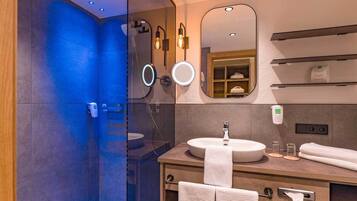 A modern bathroom with a glass shower enclosure, a round mirror, a sink with a towel, and a shelf with toiletries.