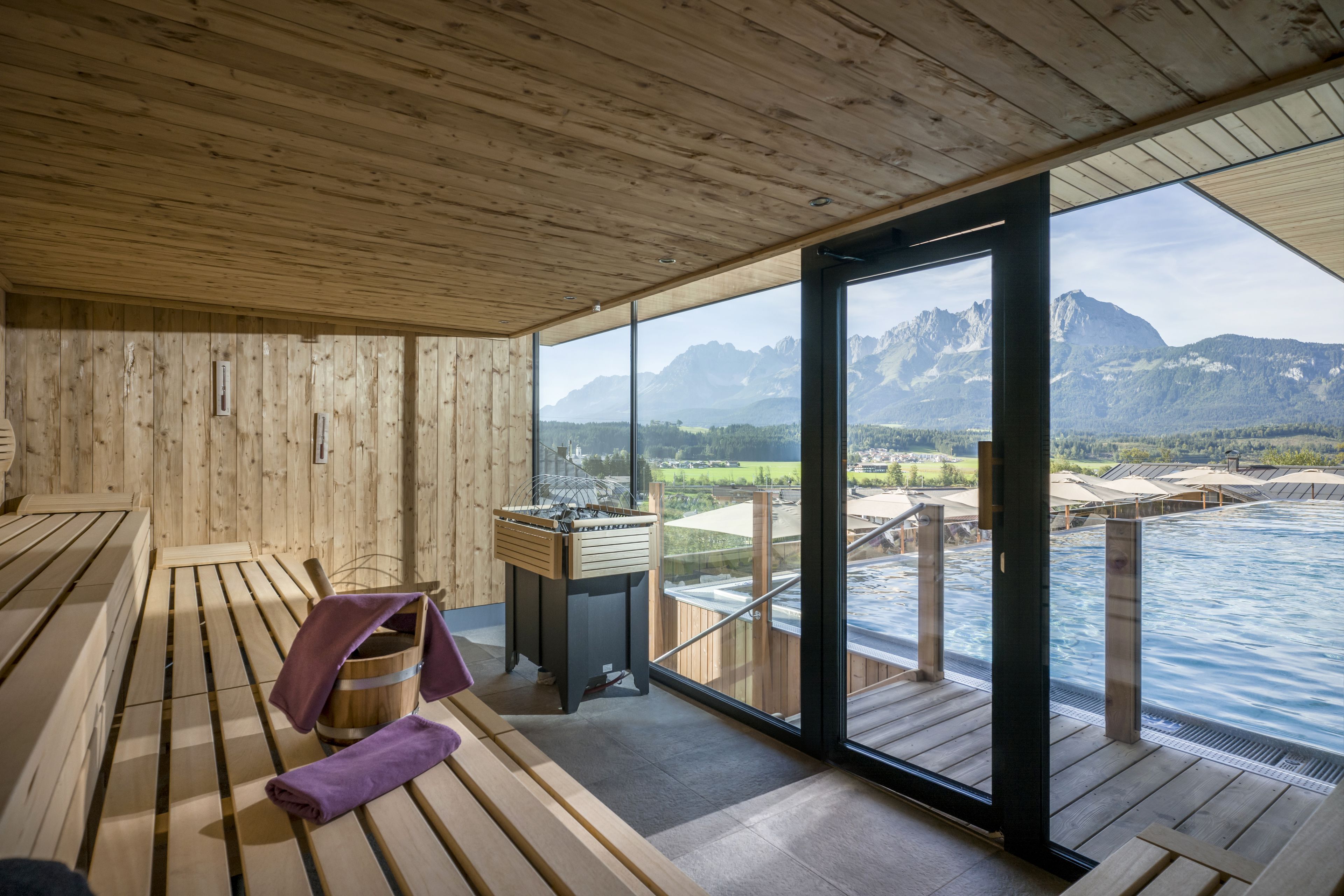 A wooden sauna room with benches and a wooden bucket, large windows offering a view of mountains and a pool.