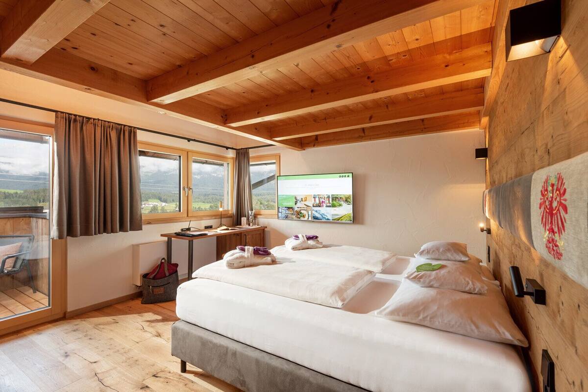 A bedroom with a large bed, wooden ceiling, a desk, and a TV on the wall.