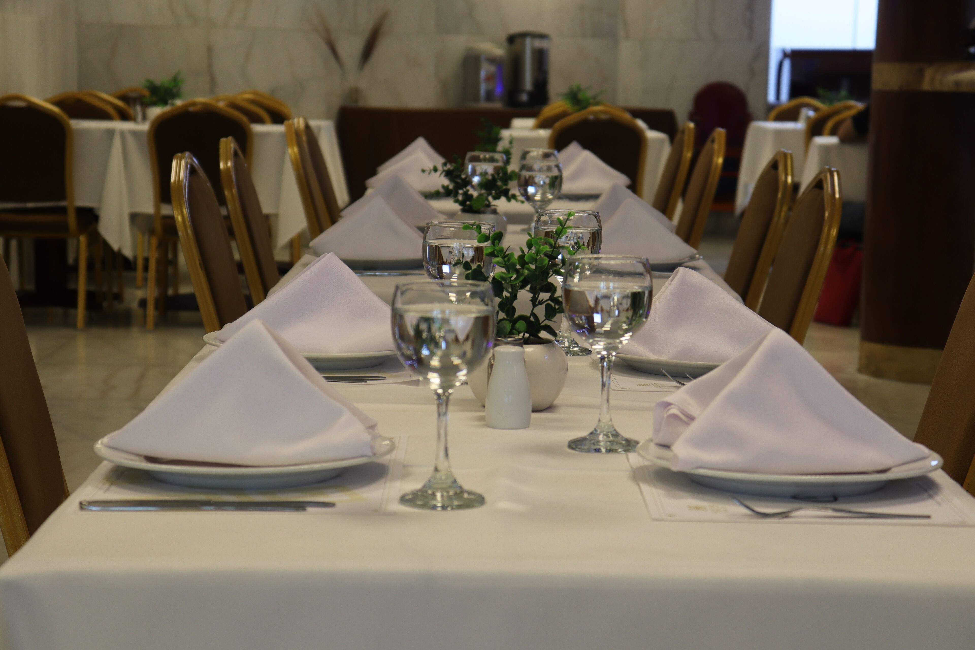 A long dining table set with white tablecloths, gold chairs, and glassware.