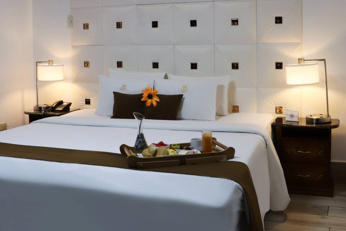Habitación de hotel con una cama grande, una mesita de noche con lámpara, una bandeja con frutas y un florero con una flor, y una pared con un diseño similar al sol.