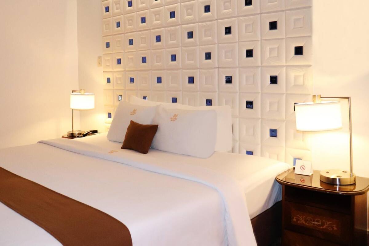 Una habitación de hotel con una cama bien hecha, dos lámparas de noche, una mesita de noche y una pared con un diseño geométrico.