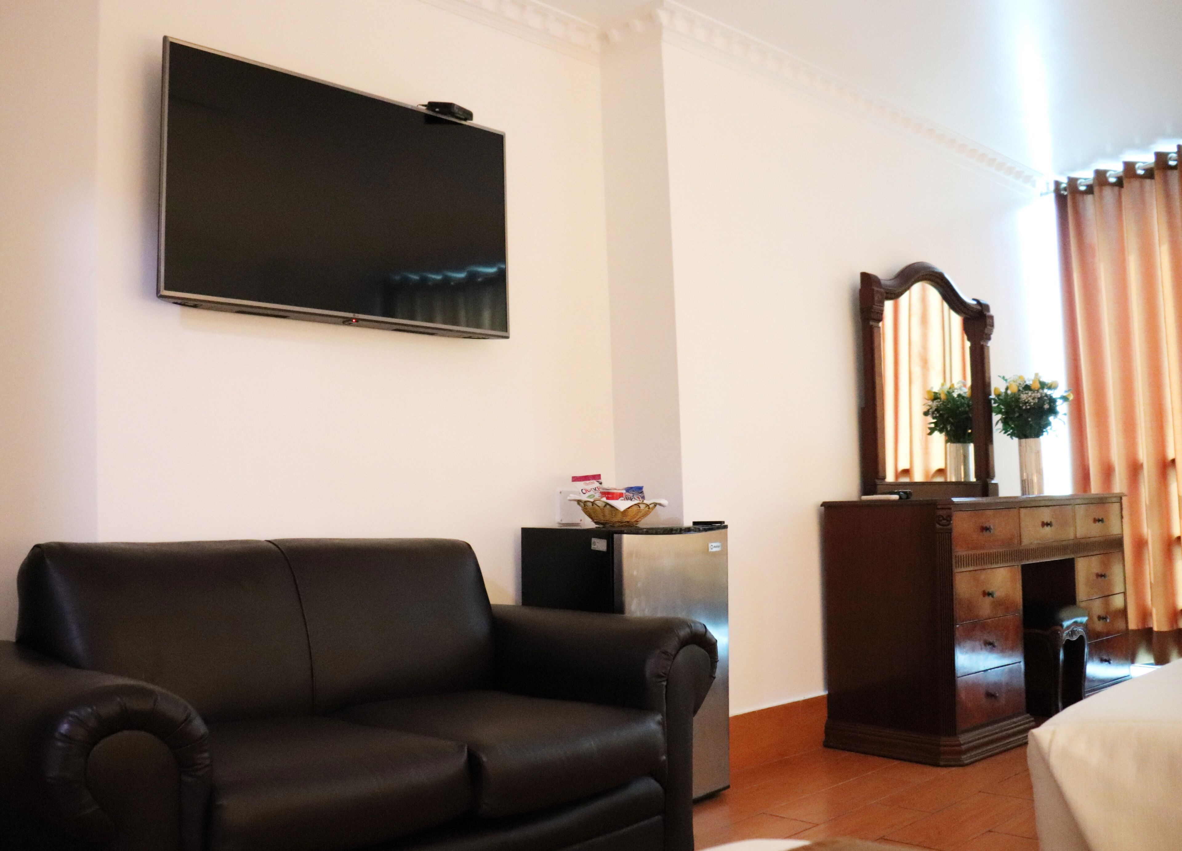 A living room with a black sofa, a flat-screen TV mounted on the wall, a small refrigerator, a wooden dresser with a mirror, and a window with curtains.